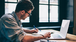 Young millennial male student searching information for homework using laptop and wifi connection indoors, man writing article in textbook for add interesting content on own website or web page