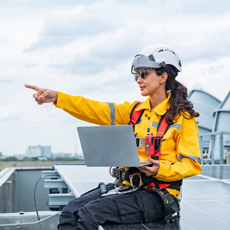 Woman in yellow safety gear smiles while examining solar panels on a rooftop and communicates via radio against an urban backdrop.