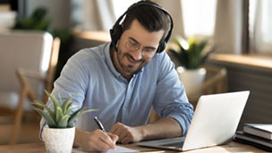 Smiling young Caucasian man in headphones glasses sit at desk work on laptop making notes. Happy millennial male in earphones watch webinar or training course or computer, study online from home.