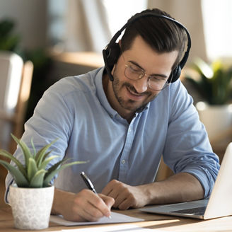 Smiling young Caucasian man in headphones glasses sit at desk work on laptop making notes. Happy millennial male in earphones watch webinar or training course or computer, study online from home.