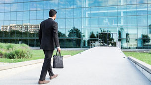 Anonymous businessman or worker in black suit with leather handbag or laptop case in his hand goes to work on the road to the entrance of a glass office building.