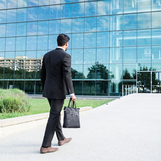 Anonymous businessman or worker in black suit with leather handbag or laptop case in his hand goes to work on the road to the entrance of a glass office building.