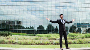 Successful happy smiling cheerful arabic businessman or worker in black suit with tie and shirt with beard standing in front of an office building with raised hands on green grass in summer day.