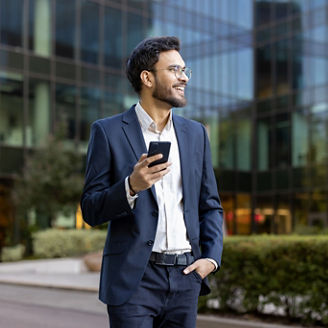 Confident young businessman in suit holding smartphone and smiling while standing outdoors near modern office building. Professional attitude, business success, and positive outlook convey success