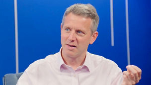 Person in a white shirt seated against a blue background, gesturing with one hand while speaking