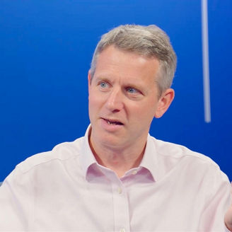 Person in a white shirt seated against a blue background, gesturing with one hand while speaking