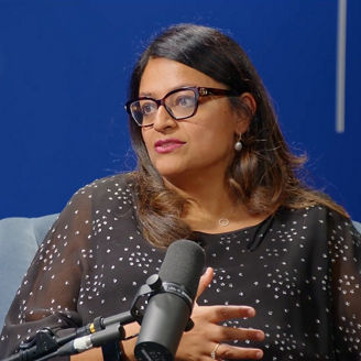 Person seated in a chair speaking into a podcast microphone. Blue studio backdrop visible behind them.