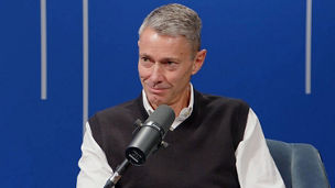 Jon Holt speaking into a microphone during a filmed discussion, seated against a blue backdrop