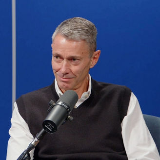 Jon Holt speaking into a microphone during a filmed discussion, seated against a blue backdrop