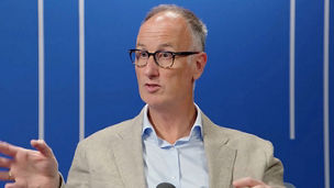 Person speaking into a microphone in a studio setting, gesturing during a discussion against a blue background with vertical light accents