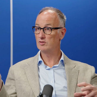 Person speaking into a microphone in a studio setting, gesturing during a discussion against a blue background with vertical light accents