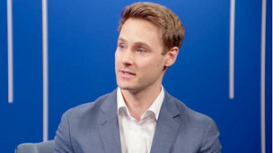 Person speaking during a studio discussion, seated against a blue background with vertical light accents