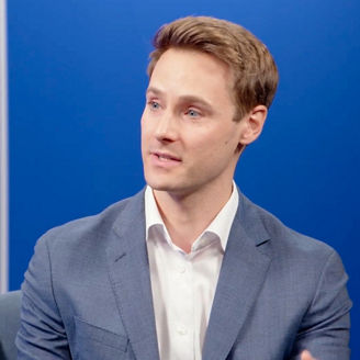 Person speaking during a studio discussion, seated against a blue background with vertical light accents