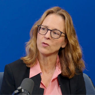 Person wearing glasses and a dark blazer speaking into a microphone during a recorded interview or panel discussion