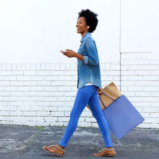 Side portrait of a smiling young woman walking with shopping bags and listening to music on smart phone