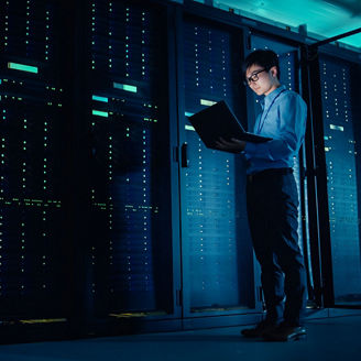 Person standing in a server room holding an open laptop, surrounded by rows of illuminated server racks in blue and purple lighting