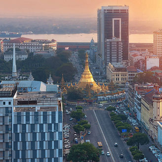 Yangon cityscape at sunset showing a central golden pagoda surrounded by tall buildings and a wide road leading toward it