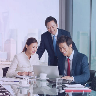 Three business professionals gathered around a conference table, looking at a laptop and documents, with a city skyline visible through large windows behind them