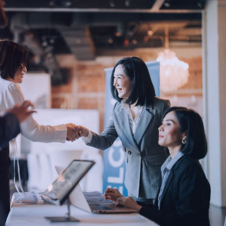 People at an event registration table, with one person standing and shaking hands while another sits at the table working on a laptop