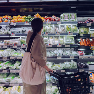 Person standing in front of a grocery store produce section with a shopping cart, looking at shelves filled with packaged vegetables and fresh produce