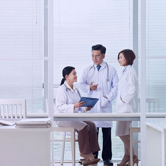 Three medical professionals in white coats standing and sitting in an office area, discussing information on a clipboard