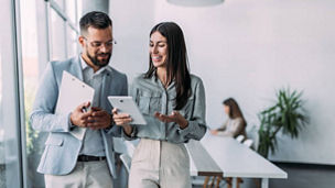 Shot of a two confident business persons talking in the work place. Two colleagues using a digital tablet while walking in a modern office. Businessman and businesswoman in meeting discussing business strategy. Business coworkers working together in the offic