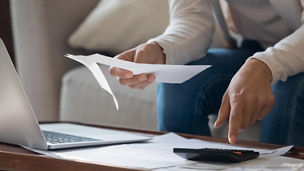 A hand is typing something on a calculator and holding a piece of paper in front of a laptop.