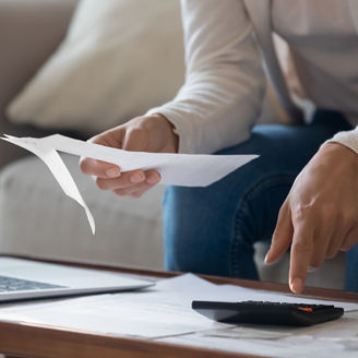 A hand is typing something on a calculator and holding a piece of paper in front of a laptop.