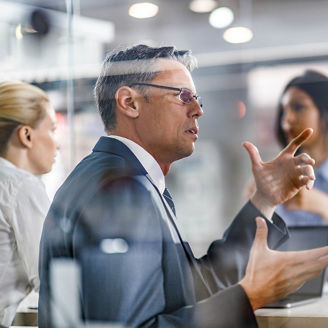 Two male entrepreneurs communicating while having a meeting with their female colleagues in the office. The view is through glass.