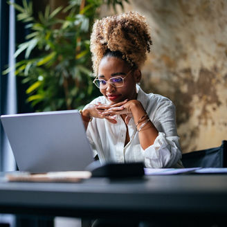 Happy African-American woman reading business report on her laptop while sitting at restaurant desk.
