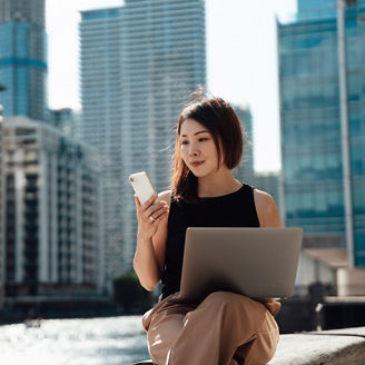 Young Asian woman using smartphone and laptop, sitting by the canal against skyscrapers in the financial district. Business, finance and investment concept.
