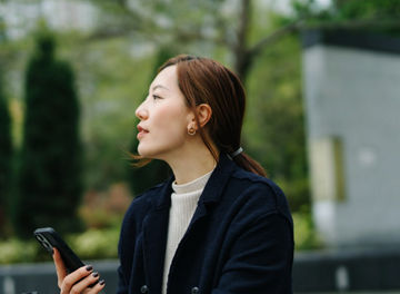 Confident young Asian businesswoman using smartphone while taking a coffee break in the middle of a work day, sitting on bench in urban office park. Fresh air to improve wellbeing