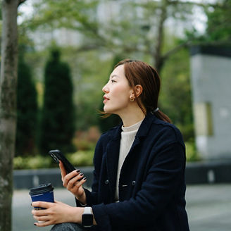 Confident young Asian businesswoman using smartphone while taking a coffee break in the middle of a work day, sitting on bench in urban office park. Fresh air to improve wellbeing