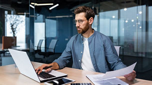 A serious young man accountant, financier, analyst, auditor sits in the office at the table. He holds documents and a pen in his hands, checks accounts, finances, types on a laptop.