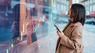 Young Asian businesswoman analysing and checking stock market over smartphone in downtown financial district. Stock exchange market trading board in background.