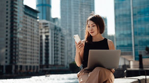 Young Asian woman using smartphone and laptop, sitting by the canal against skyscrapers in the financial district. Business, finance and investment concept.