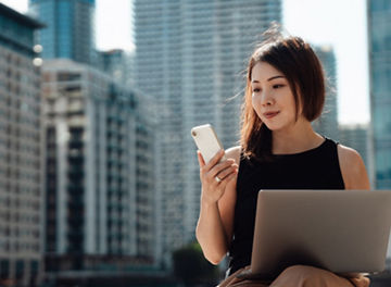 Young Asian woman using smartphone and laptop, sitting by the canal against skyscrapers in the financial district. Business, finance and investment concept.