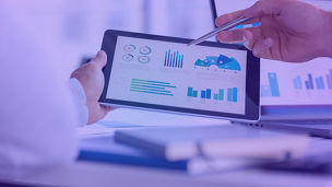 Two business men meeting and looking at financial data, charts and graphs on a digital tablet and a laptop computer. There are documents and paperwork on the table. One man is pointing at the screen