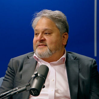 “Person speaking into a microphone in a studio setting, seated against a blue background with vertical light accents