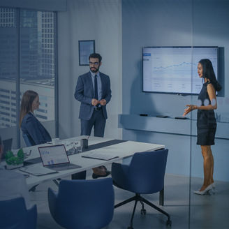 Konferenzraum mit Glaswänden: mehrere Personen sitzen an einem Tisch, während eine Person an einem Wandbildschirm mit Diagrammen präsentiert; Laptops auf dem Tisch, Blick auf Stadtgebäude im Hintergrund.