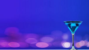 Das Bild zeigt ein elegantes Cocktailglas, das mit einem blauen Getränk gefüllt ist. Der Hintergrund ist ein lebendiger Farbverlauf aus Blau- und Violetttönen, der eine stimmungsvolle und festliche Atmosphäre schafft. Das Glas steht rechts im Bild und hebt sich durch seine klare Form und Farbe vom Hintergrund ab.