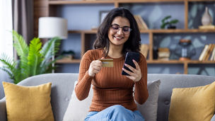 Smiling young Indian girl sitting on sofa at home, using credit card and mobile phone.