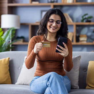 Smiling young Indian girl sitting on sofa at home, using credit card and mobile phone.