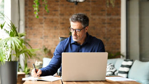 Busy mature middle aged professional businessman executive using laptop at work writing notes. 50 years old business man company manager investor working on computer technology sitting at office desk.