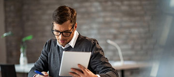 Male entrepreneur taking notes while going through paperwork and using touchpad at work. 