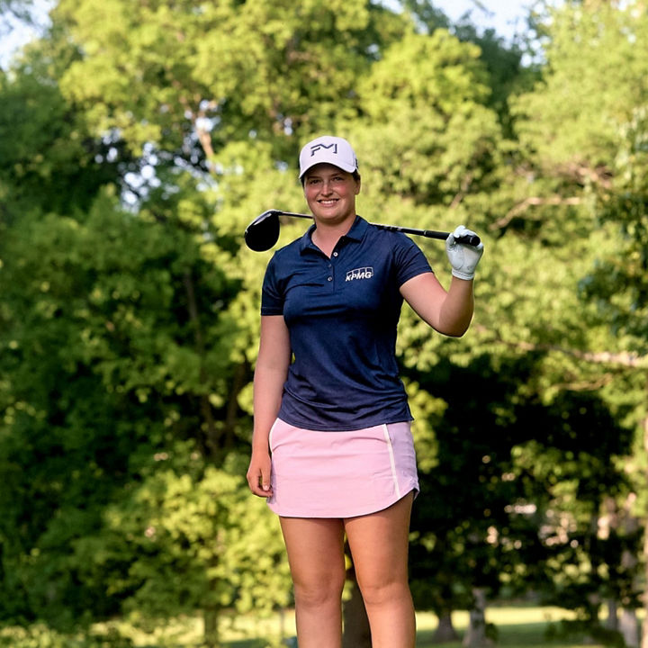 Lottie Woad, LPGA Professional Golfer standing on a golf course holding a club, with trees and greenery in the background