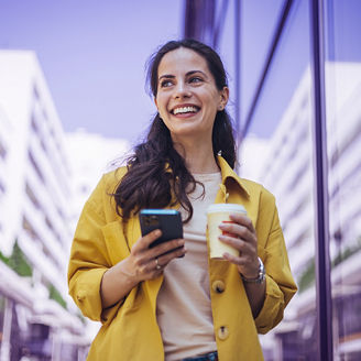 Modern young woman walking on the city street texting and holding cup of coffee. Smiling woman using mobile phone while having a coffee. Business woman texting on her phone 