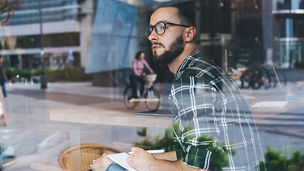 Through glass side view of serious bearded black haired man in sunglasses and checkered shirt sitting in cafe with notebook and looking out window; Shutterstock ID 1761071057; Client: Wafae Bouari; job: AI blog 3 - transparantie; Team: Assurance; Image in line with KPMG guidelines?: Yes