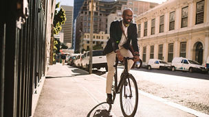 Young smiling businessman going to work by bike. Man in suit riding bicycle on sidewalk in morning.