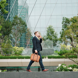 Side view of businessman calling manager about marketing report and walking at modern building in green eco city. Leader going to workplace while talking to his team by using phone. Lifestyle. Urbane.